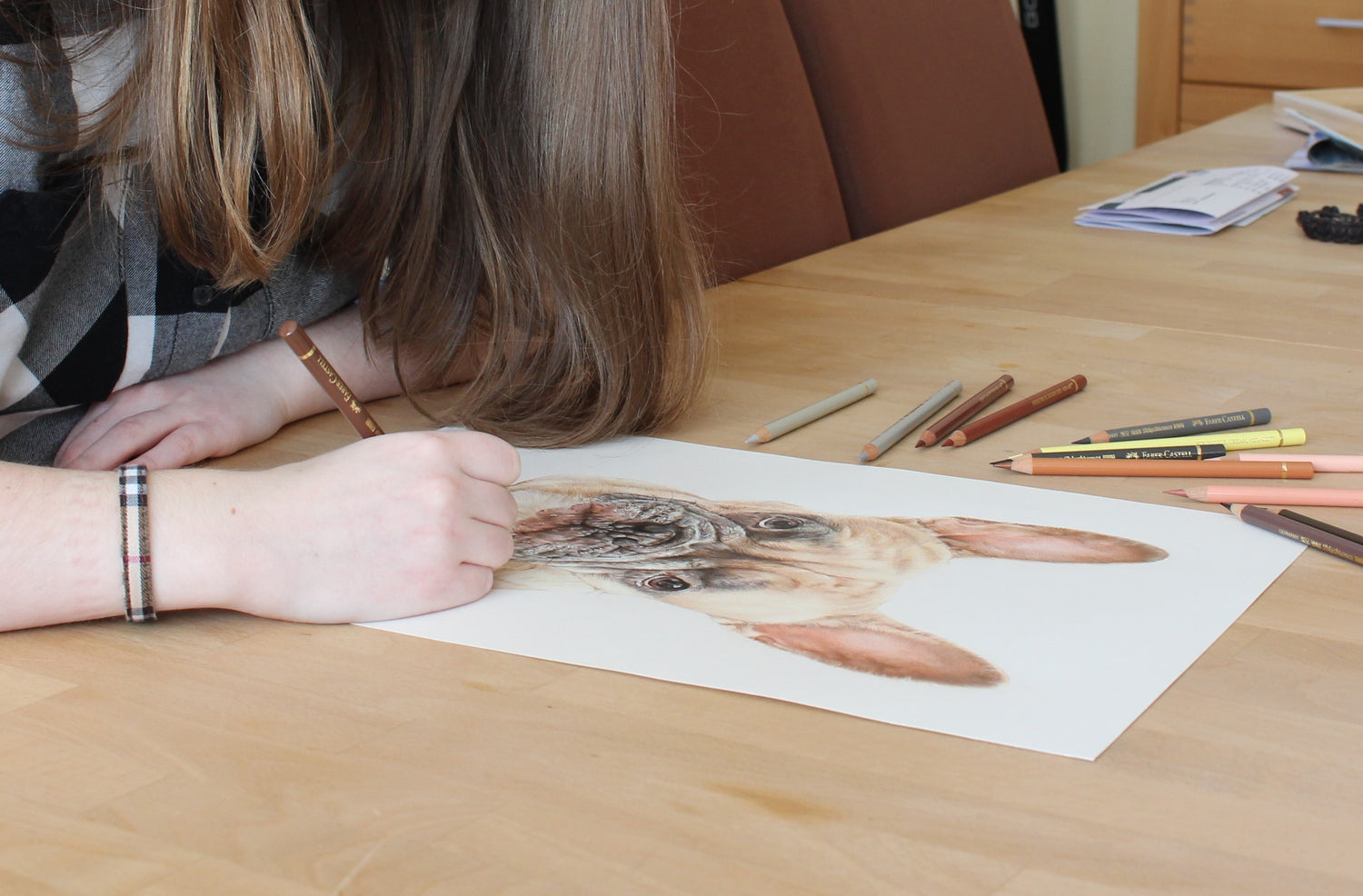 Papier mit Zeichnung einer französischen Bulldogge liegt auf dem Tisch, Hand hält einen braunen Stift und liegt auf dem Blatt auf